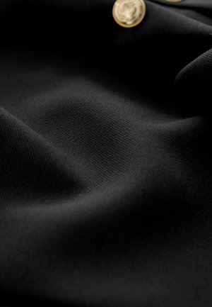 Schwarzer Stoff mit einer glatten, strukturierten Oberfläche, ausgestattet mit goldfarbigen Knöpfen mit einem erhöhten Emblem. Das Material ist weich und leicht elastisch.
