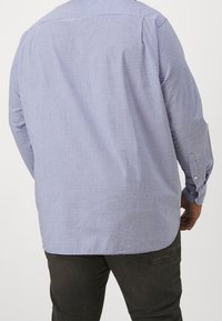 Man in een lichtblauw en wit klein geblokt overhemd met lange mouwen en donkere jeans, staand met de rug naar de camera.