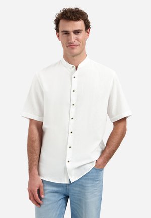 Jonge man met krullend haar, gekleed in een wit overhemd met korte mouwen en een lichtblauwe spijkerbroek, staand met één hand in zijn zak.