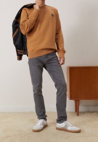 Hnědá mikina s malým logem, šedé džíny a bílé tenisky s gumovými podrážkami. Model drží černou bomber bundu. Neutrální pozadí.