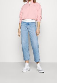 Světlounce růžový pulovr na půl zip s texturovaným povrchem, spárovaný se světle modrými zkrácenými džínami. Modelka má na nohou bílé tenisky s černými doplňky.