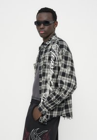 Πουκάμισο με ασπρόμαυρο καρό, φθαρμένο τελείωμα, μακριά μανίκια και λεπτομέρεια με κείμενο στο πλάι, συνδυασμένο με σκούρο παντελόνι με κόκκινο μοτίβο. Μαύρα γυαλιά ηλίου.