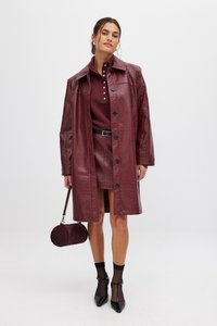 Cappotto in ecopelle bordeaux con chiusura a bottoni, finitura testurizzata, abbinato a una gonna coordinata e calze nere alla caviglia, mentre tiene una borsa rotonda.