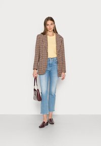 Blazer xadrez em castanho e creme, suéter amarelo, jeans azul claro, mocassins bordôs e uma pequena mala bordô com um padrão texturizado.