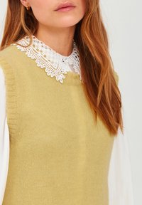 Light yellow knit vest with a ribbed neckline layered over a white blouse featuring a lacy collar detail. Soft texture, simple design.