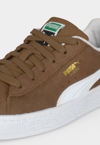 Καφέ σουέντ αθλητικό παπούτσι Puma με λευκή ρίγα, λευκή σόλα, καφέ κορδόνια και πράσινο λογότυπο Puma στη γλώσσα.