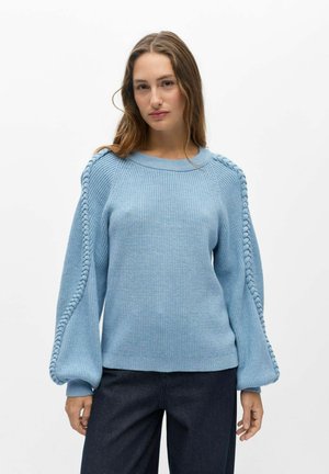 Jeune femme portant un pull en maille bleu clair avec des détails tressés sur les manches et un pantalon foncé, debout devant un fond blanc uni.