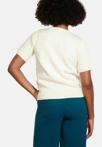 Crème kleurige gebreide trui met korte mouwen, ribbelboord en halslijn, gedragen met teal broek. De trui heeft decoratieve details op de achterkant.