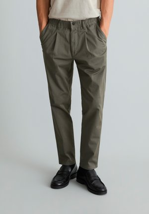Man draagt olijfgroene geplooide broek, wit t-shirt ingestopt, handen in de zakken en zwarte leren loafers op een lichtgrijze vloer.