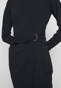 Robe noire à manches longues avec détail de tissu froncé et un anneau métallique à la taille, portée par une personne les mains derrière le dos.