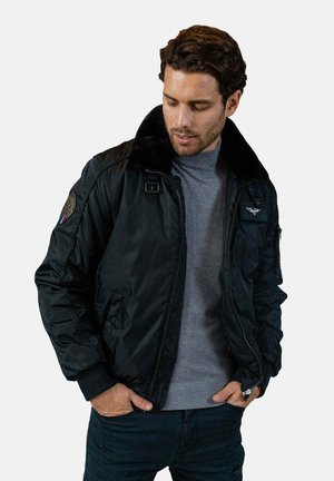 Veste bomber noire avec un col en fausse fourrure, fermeture éclair et des patchs brodés sur les manches. Matière lisse et poignets côtelés.