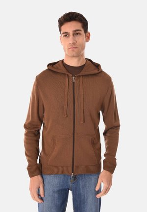 Uomo che indossa un maglione marrone con cappuccio e cerniera, con tasche frontali e jeans blu, in piedi davanti a uno sfondo chiaro e uniforme.