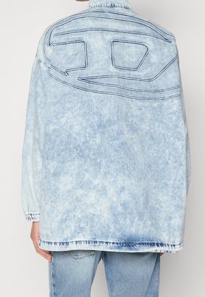 Person iført lyseblå acid-wash denimjakke med syet abstrakt design på ryggen, kombineret med blå jeans, mod en ensfarvet baggrund.