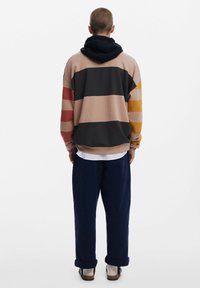 Sudadera con capucha en beige con rayas horizontales en negro, rojo y amarillo. Corte holgado, tela texturizada, con una capucha forrada en navy y puños acanalados.