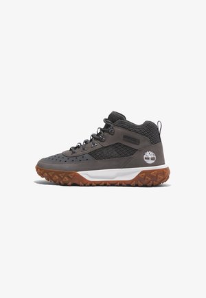 Scarpa da trekking a metà altezza con tomaia in suede grigio e mesh, suola in gomma con tacchetti triangolari e intersuola bianca con accento del logo.