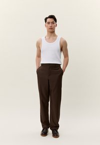 Mladi moški stoji pokončno z rokami v žepih, oblečen v belo majico brez rokavov, rjave hlače in temne usnjene čevlje.