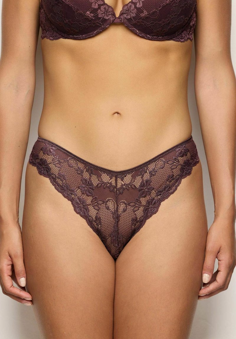 Koronkowy string w głębokim fiolecie z kwiatowymi wzorami, z falbaniastym paskiem i przezroczystymi sekcjami, uwydatniającym skomplikowane detale i fakturę.