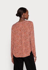 Blusa de manga longa em cor ferrugem com padrão floral, apresentando uma bainha arredondada e um corte descontraído. Textura suave com mangas ligeiramente largas.