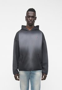 Νεαρός άνδρας που φοράει μαύρο φούτερ με βαθμιδωτό χρωματισμό, εμπρόσθιες τσέπες και ανοιχτό μπλε τζιν, στέκεται μπροστά σε λευκό φόντο.