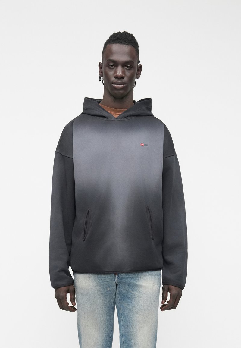 Νεαρός άνδρας που φοράει μαύρο φούτερ με βαθμιδωτό χρωματισμό, εμπρόσθιες τσέπες και ανοιχτό μπλε τζιν, στέκεται μπροστά σε λευκό φόντο.