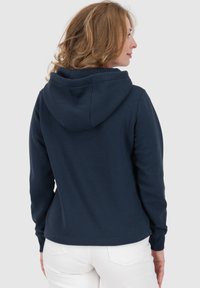 Felpa blu navy realizzata in tessuto morbido, con cappuccio, maniche lunghe e design aderente. Pantaloni bianchi sono visibili in basso.
