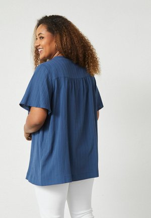 Blå kortärmad topp med ett texturerat mönster, avslappnad passform och en rynkad ryggstycke. Vita byxor fulländar outfiten.