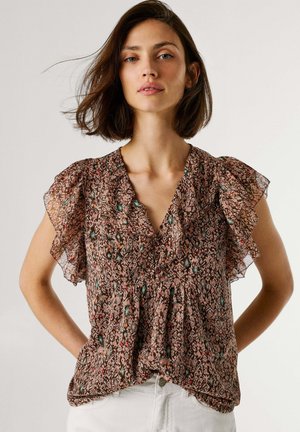 Kobieta o brązowych włosach do ramion, ubrana w wzorzystą, krótka bluzkę z falbankami i białe spodnie, pozująca z rękami na biodrach.