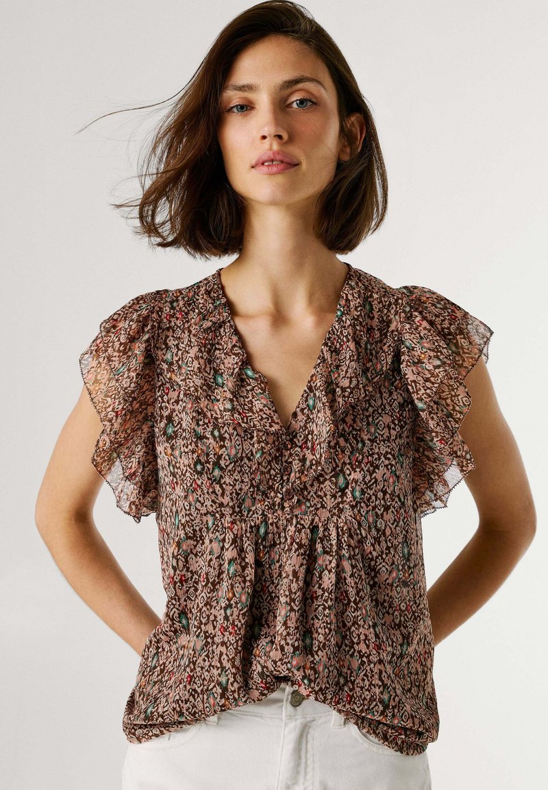 Frau mit schulterlangem braunem Haar, die eine gemusterte, kurzärmelige Bluse mit Rüschen und weiße Hose trägt, posiert mit den Händen in der Hüfte.