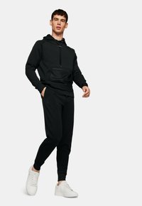 Sweatshirt noir avec fermeture éclair quarts et poche avant, associé à un jogging noir assorti et des baskets blanches. Tissu lisse, coupe décontractée.