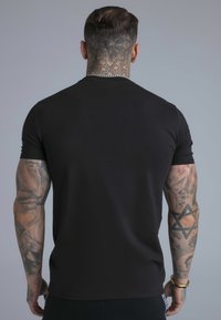 Musta vartalonmyötäinen lyhythihainen t-paita, valmistettu sileästä materiaalista. Näkyvät tatuoinnit käsivarsissa ja kaulalla, minimalistinen muotoilu ilman grafiikkaa.