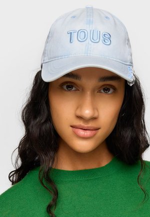 Mujer con cabello largo y oscuro lleva una gorra de béisbol azul claro con "TOUS" bordado, combinada con un suéter verde, fondo neutro.