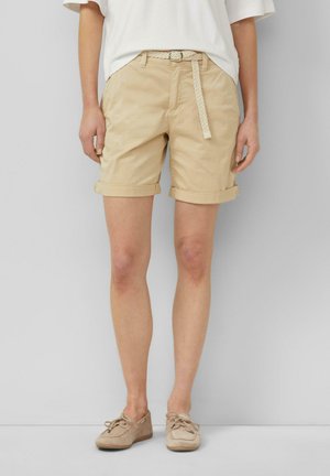 Persoon die beige opgerolde korte broek draagt met een geweven riem, een wit topje en beige veterschoenen, staande tegen een effen achtergrond.