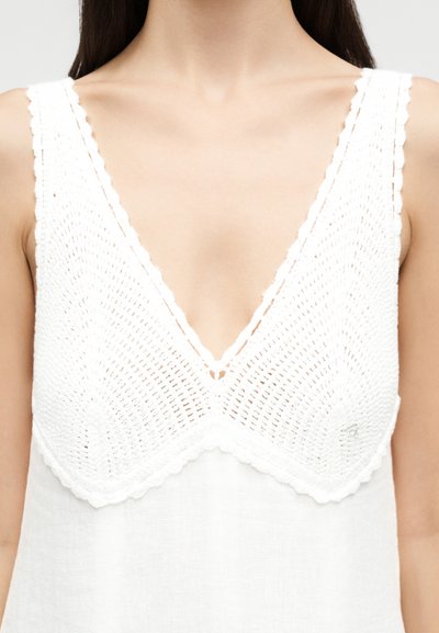 Nő ujjatlan fehér horgolt felsőben, V-nyakú, ami láttatja a mellkas felső részét és a vállakat, semleges hátteret használva.