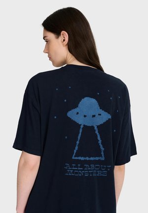 Femme aux longs cheveux foncés porte un t-shirt noir oversize avec un OVNI pixelisé bleu et le texte "ALL ABOUT MONSTERS" dans le dos.