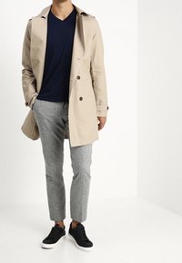 Beige trenchcoat met dubbele rij knopen, marineblauw V-hals shirt, grijze taps toelopende broek en zwarte sneakers met witte zolen.
