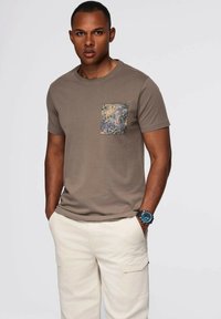 Hnědé tričko s vzorovanou náprsní kapsou v tlumených barvách. Má krátké rukávy a kulatý výstřih. Nosí se s krémovými kalhotami.