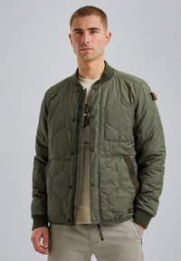 Giacca bomber trapuntata verde oliva con bottoni a pressione, polsini a coste, tasche laterali e texture imbottita. Sotto, è visibile una camicia di colore chiaro.