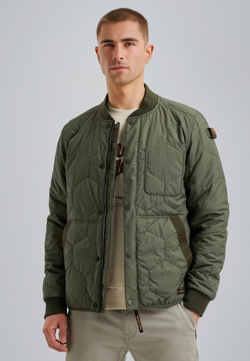 Giacca bomber trapuntata verde oliva con bottoni a pressione, polsini a coste, tasche laterali e texture imbottita. Sotto, è visibile una camicia di colore chiaro.