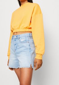 Pullover giallo tagliato con maniche lunghe e fondo elastico, abbinato a pantaloni corti in denim azzurro chiaro con orli sfilacciati e leggeri segni di usura.