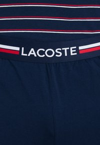 Tengerészkék pamut rövidnadrág fehér szegéllyel, piros és kék csíkokkal, valamint "LACOSTE" logóval. Az anyag puha és sima textúrájú.