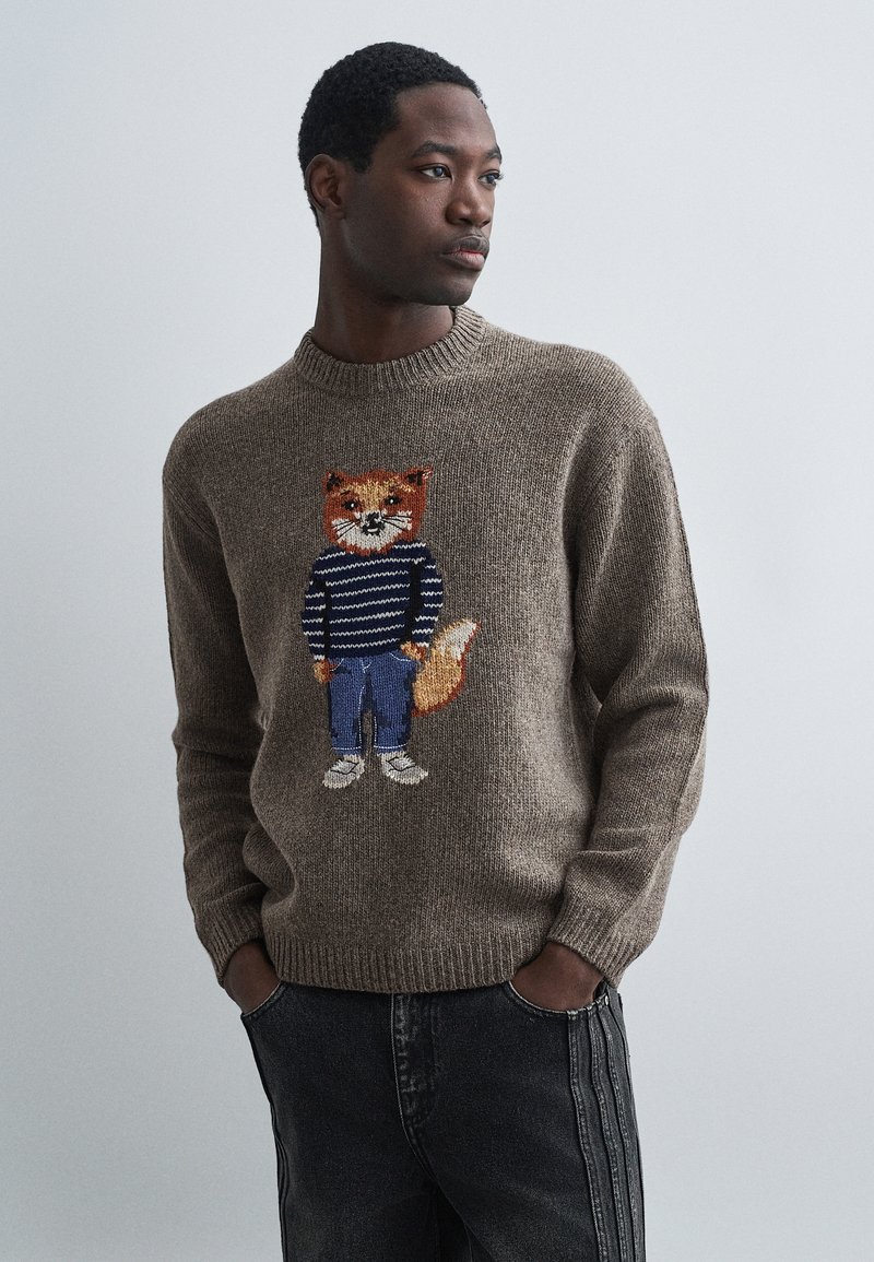 Vīrietis, kurš valkā brūnu adītu džemperi ar lapsu uz svītrainas krekla un džinsu dizaina, stāv ar rokām kabatās uz vienkrāsaina fona.