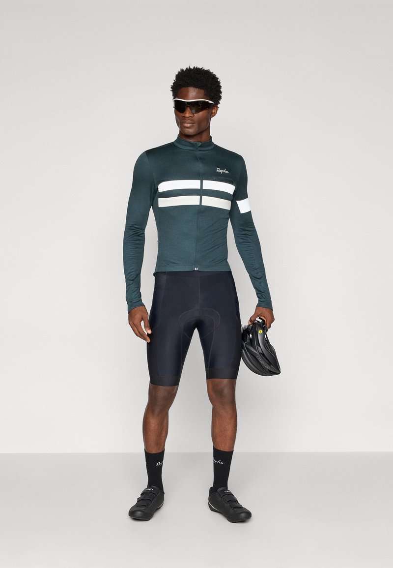Dark green cycling jersey with white stripes, zippered front, long sleeves, paired with black shorts and black cycling shoes. Holding a helmet.