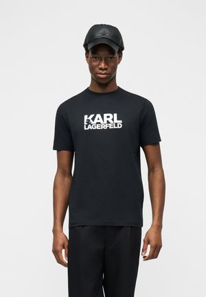 Moški v črni usnjeni kapi, črni majici s kratkimi rokavi z belim napisom »KARL LAGERFELD« in črnih hlačah stoji pred enobarvnim svetlim ozadjem.