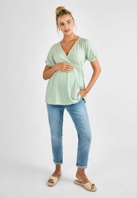 Világos zöld, textúrált kismama felső V-nyakkal és rövid puffos ujjakkal, világoskék, feltekert farmernadrággal és bézs csúsztatós szandálokkal párosítva.