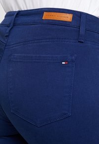 Jeans de mezclilla azul oscuro con un parche de cuero con el logo de Tommy Hilfiger en la cintura, que cuentan con un bolsillo lateral con un pequeño acento en rojo, blanco y azul.