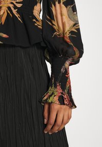 Primo piano di una mano che indossa una blusa nera a fiori con polsini arricciati e una gonna nera plissettata su uno sfondo bianco.