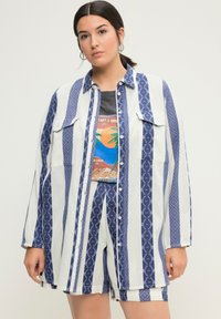 Camicia a righe blu e bianche con motivi geometrici, con bottoni frontalmente e due tasche sul petto, indossata sopra una maglietta grafica e pantaloni corti abbinati.