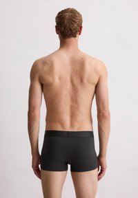 Shorts de boxeo negros que lleva un modelo masculino, mostrando un diseño ajustado y una textura suave. La espalda del modelo es visible, resaltando el tono de piel y la forma del cuerpo.