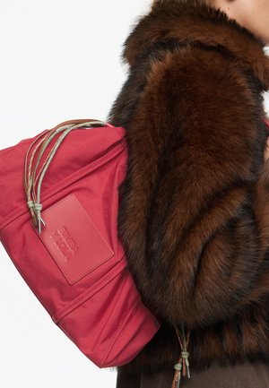 Bolso de hombro de tela roja con cierre de cordón y detalles de cuero, que presenta una placa con el logo. El bolso se lleva con un abrigo de piel sintética marrón.