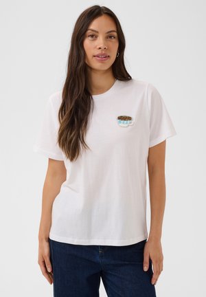 Femeie purtând un tricou alb cu o cană mică de cafea brodată lângă piept, asortat cu blugi de culoare albastru închis.